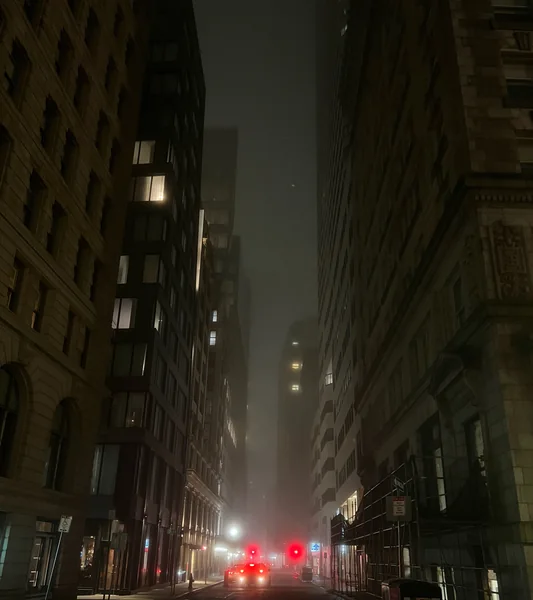 A narrow street between tall buildings ends in a glow of red traffic signals.