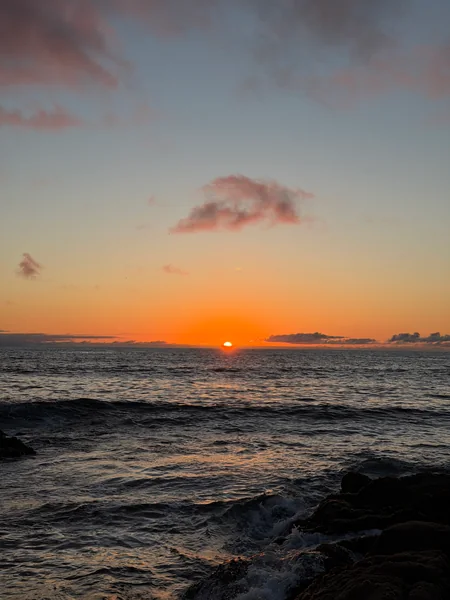 The sun rests near the horizon above waves and dark shoreline rocks.