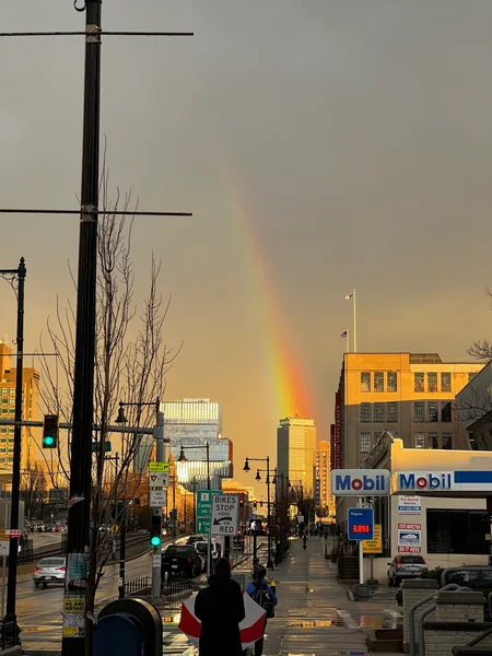 A rainbow appears above a neighborhood street, pedestrians and roadside signs.