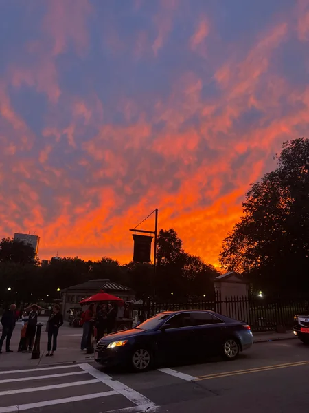 Vivid orange clouds rise above a street corner, pedestrians and a passing car.