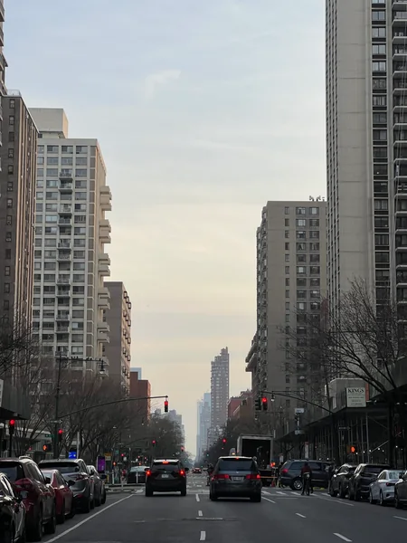 Traffic and buildings recede down a city avenue under a pale sky.