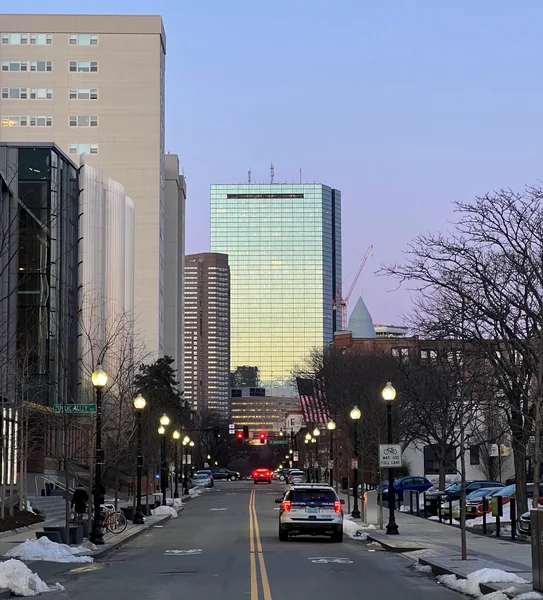 A city avenue with traces of snow leads toward glass towers under a blue sky.